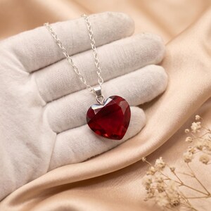 May include: A faceted deep red heart-shaped pendant suspended from a simple silver-toned metal chain. The pendant features a polished, multi-faceted surface that reflects light, creating a brilliant appearance against the dark crimson gemstone.
