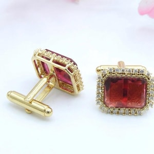 May include: Gold-coloured cufflinks featuring rectangular, ruby-red centre stones. Each cufflink is framed by a border of small, clear crystals. The cufflinks have a classic design, suitable for formal occasions.