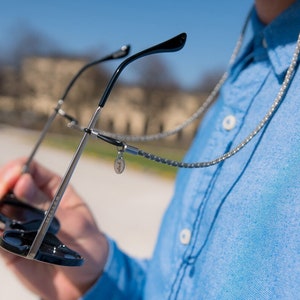 May include: Sunglasses with a silver frame and black temples are held up by a silver chain. The chain is attached to the glasses and hangs down. A small, round charm dangles from the chain.