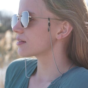 Puede incluir: Una mujer con gafas de sol con una cadena plateada unida a las monturas. La cadena cuelga alrededor de su cuello. Lleva una camisa turquesa y tiene el pelo largo y rubio.
