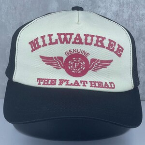 May include: A black and off-white trucker hat featuring a red graphic print on the front panel. The text reads MILWAUKEE, GENUINE, and THE FLAT HEAD, with a central circular emblem containing a letter F and stylized wings on either side.