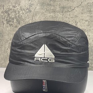 May include: A black five-panel cap made of a lightweight, crinkled nylon material. The front features an embroidered white triangular logo with the text ACG and a small Nike swoosh. The cap has a flat brim and a structured crown design.