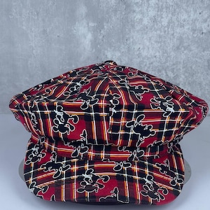 May include: A newsboy cap featuring a red, black, and yellow plaid pattern with a repeating black silhouette graphic of a cartoon mouse character. The hat has a rounded, multi-paneled crown and a short, stiff brim extending from the front.