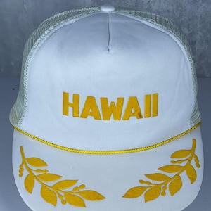 Può includere: Un cappellino trucker bianco con retro in rete e pannello frontale solido. La parola HAWAII è ricamata in lettere maiuscole gialle sul davanti. Un sottile cordoncino giallo corre lungo la base della corona e due rametti di foglie gialle sono ricamati sulla visiera.