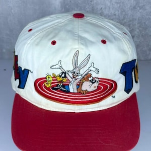 May include: A white baseball cap with a red brim and a red button on top. The front features an embroidered graphic of Bugs Bunny, Daffy Duck, Tweety Bird, and the Tasmanian Devil emerging from a red concentric circle target design.