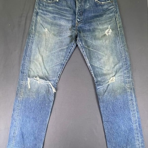 May include: A pair of blue denim jeans featuring a distressed finish with fading, whiskering, and frayed holes at the knees and pockets. The jeans have a button fly closure, a classic five-pocket design, and a straight leg cut with worn hems.