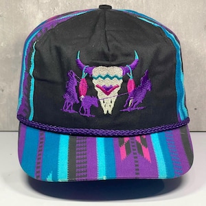 Può includere: Un cappellino da baseball nero con un ricamo centrale raffigurante un teschio di toro bianco con accenti viola e turchesi. Il cappellino presenta un cordino intrecciato viola alla base della corona e una visiera con un motivo colorato.