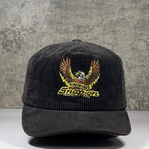 Pode incluir: Um boné de beisebol de veludo cotelê preto com um gráfico bordado de uma águia com asas abertas acima do texto amarelo "Snap-on" e o texto menor "SINCE 1920" centralizado no painel frontal do boné com uma coroa estruturada.