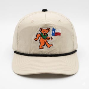Puede incluir: Gorra de béisbol beige con un detalle de cuerda negra y un botón negro en la parte superior. La gorra presenta un oso naranja bordado con un cuello verde que sostiene una bandera de Texas. El sombrero es de estilo clásico.