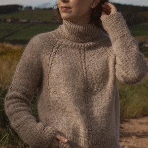 May include: A brown turtleneck sweater with a cable knit pattern. The sweater is worn by a person standing in a field.