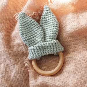 May include: A light blue crocheted bunny rabbit teether with a wooden ring. The teether is on a pink and brown textured surface.