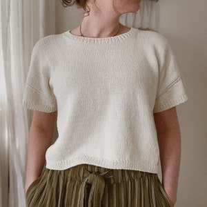Può includere: Top in maglia color crema a maniche corte con scollo tondo. Il top ha un design semplice e classico. La modella indossa pantaloni a righe verdi e bianche.