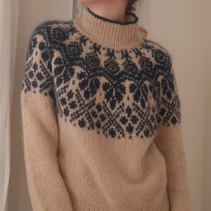 Op de afbeelding: Een beige coltrui met een marineblauw geometrisch patroon over de bovenborst en schouders. De trui heeft lange mouwen en een geribbelde zoom en manchetten. De halslijn en manchetten zijn afgewerkt met marineblauw.