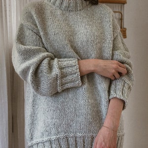 Puede incluir: Un suéter gris claro de gran tamaño con cuello y puños de canalé. El suéter está hecho de un hilo suave y texturizado.