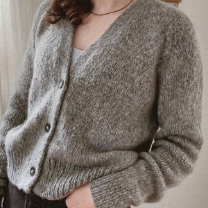 Puede incluir: Un cárdigan gris con botones y cuello en V. El suéter presenta un tejido texturizado y mangas largas. Los botones son oscuros y el estilo es casual. El color neutro lo hace versátil. El cárdigan es cómodo.