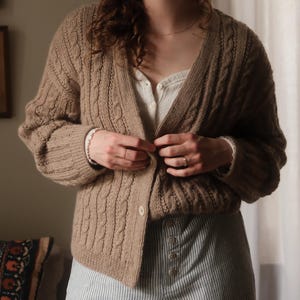 May include: A tan cable-knit cardigan with a V-neckline and front button closure, worn over a white button-down shirt and light blue and white pinstriped trousers. The cardigan features vertical cable patterns and ribbed cuffs.