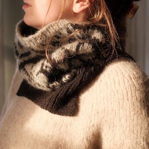 May include: A cozy, patterned cowl scarf in shades of gray, black, and cream. The scarf is made of a soft, textured knit material and is designed to be worn around the neck. The cowl is a warm and stylish accessory for cold weather.
