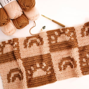 May include: A crocheted blanket with a geometric pattern in brown and pink yarn. The blanket is made of squares with a repeating design of arches. There are balls of yarn in brown and pink in the top left corner of the image.