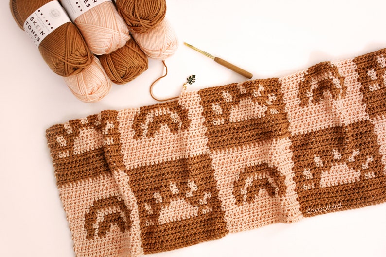 May include: A crocheted blanket with a geometric pattern in brown and pink yarn. The blanket is made of squares with a repeating design of arches. There are balls of yarn in brown and pink in the top left corner of the image.