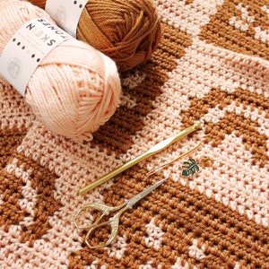 May include: A crocheted blanket with a chevron pattern in brown and pink yarn. The blanket is laid out flat on a white surface. There are two balls of yarn, a crochet hook, and a pair of scissors on the blanket. The yarn balls have labels that say "Jones Skein".