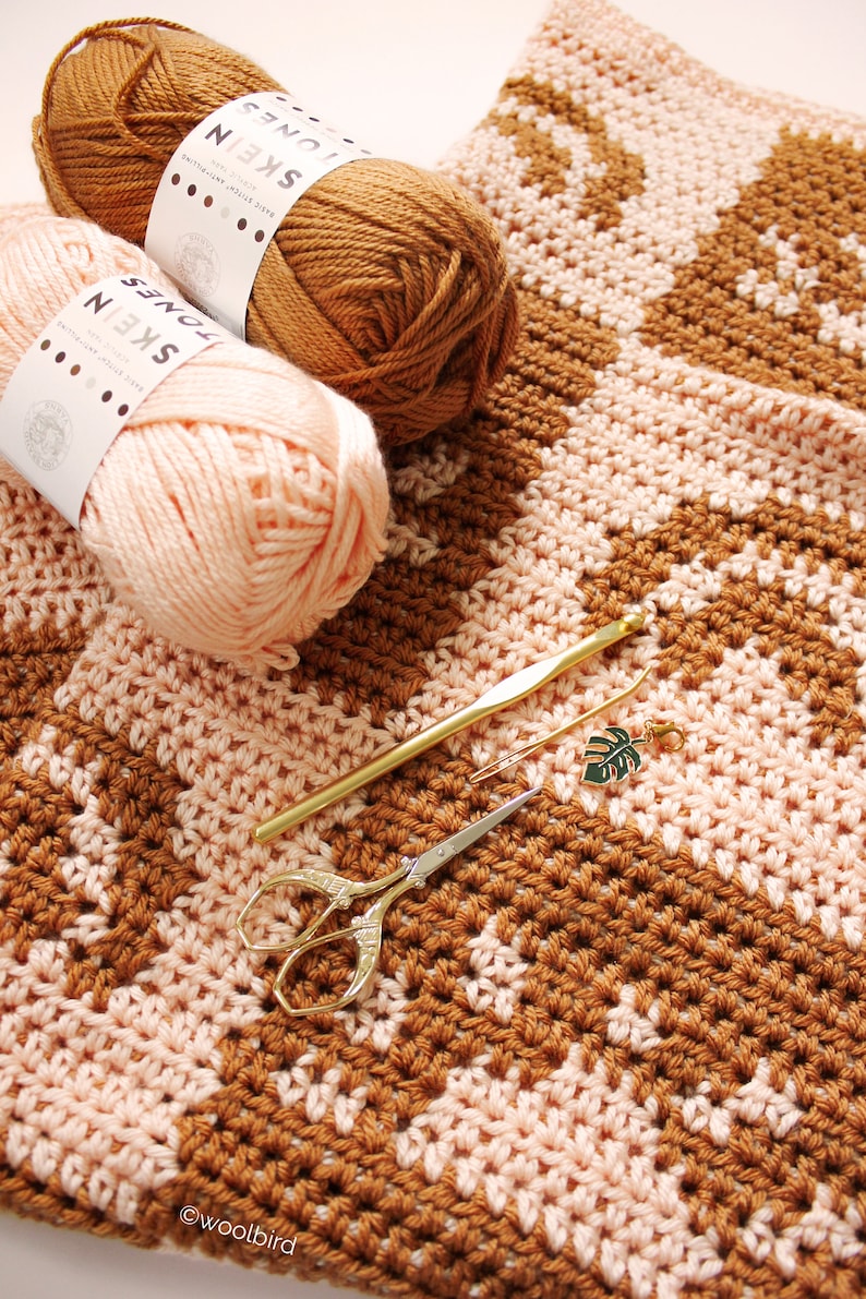 May include: A crocheted blanket with a chevron pattern in brown and pink yarn. The blanket is laid out flat on a white surface. There are two balls of yarn, a crochet hook, and a pair of scissors on the blanket. The yarn balls have labels that say "Jones Skein".