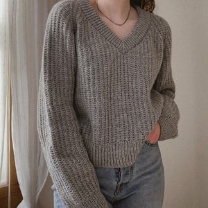 May include: A gray, ribbed knit sweater with a V-neck and long sleeves. The sweater is paired with light blue denim jeans. The overall style is casual and comfortable, suitable for everyday wear. The sweater's texture is prominent.