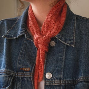 May include: A coral-coloured knitted scarf tied in a knot, worn over a denim jacket. The scarf features an openwork design. The denim jacket has silver buttons and a classic, faded blue wash.