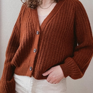 May include: A rust-colored knitted cardigan with a v-neck and button closure. The cardigan has a ribbed texture and long sleeves.