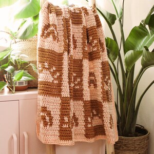 May include: A crocheted blanket with a geometric pattern in shades of brown and pink. The blanket is hanging on a wooden ladder against a pink cabinet.