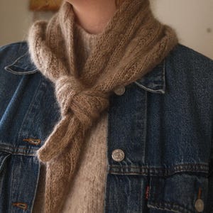 Puede incluir: Bufanda de punto marrón claro anudada. La bufanda se lleva sobre un suéter beige y una chaqueta vaquera azul. La bufanda parece estar hecha de un hilo suave y texturizado, y es de color neutro.