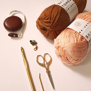 May include: Two skeins of yarn, one brown and one peach, with the label "Skein Tones" on each.  The yarn is next to a brown tape measure, a gold crochet hook, a gold tapestry needle, and a pair of gold scissors.