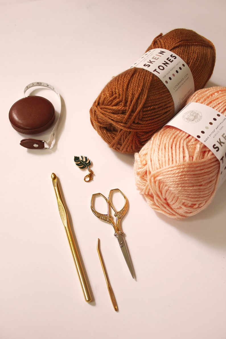 May include: Two skeins of yarn, one brown and one peach, with the label "Skein Tones" on each.  The yarn is next to a brown tape measure, a gold crochet hook, a gold tapestry needle, and a pair of gold scissors.