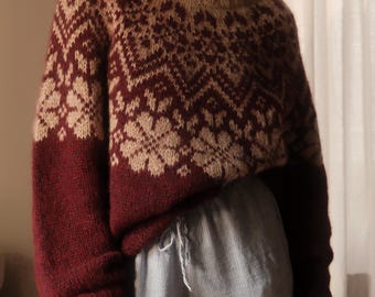 May include: A long-sleeved knit sweater featuring a deep burgundy and beige color scheme with a repeating floral and geometric pattern across the chest and shoulders, finished with a ribbed crew neckline and ribbed cuffs.
