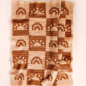 May include: A crocheted baby blanket with a checkered pattern featuring rainbows, suns, and stars in shades of brown and pink. The blanket is folded and lying on a white surface. The text "woolbird" is visible in the lower right corner.