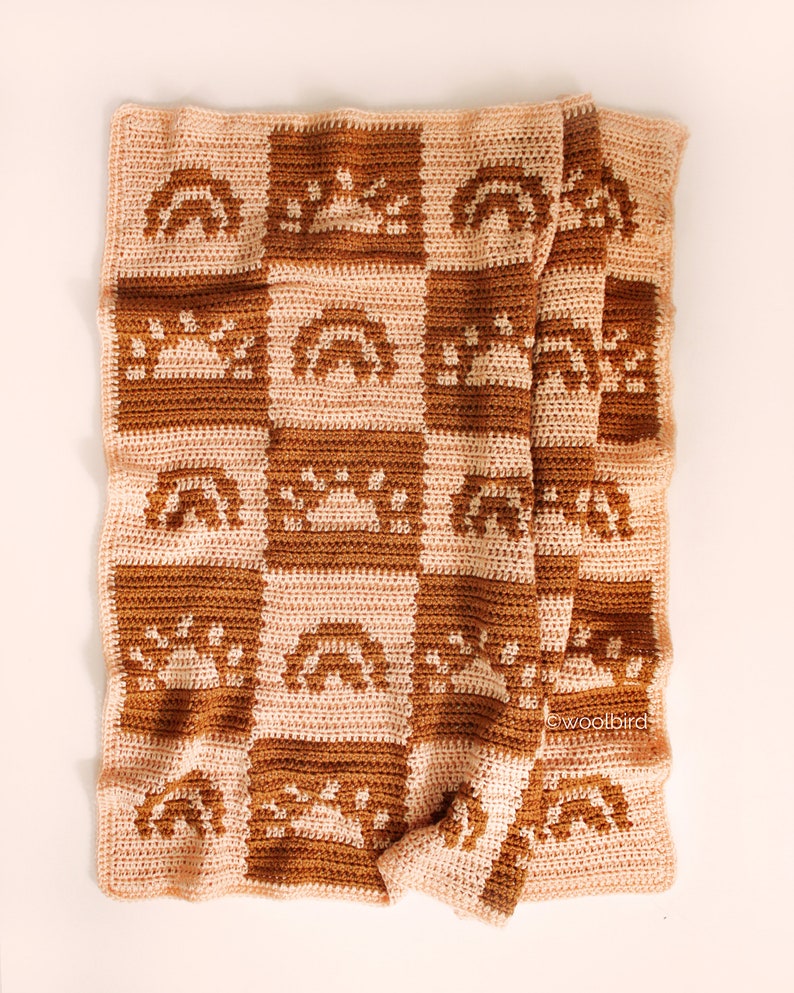 May include: A crocheted baby blanket with a checkered pattern featuring rainbows, suns, and stars in shades of brown and pink. The blanket is folded and lying on a white surface. The text "woolbird" is visible in the lower right corner.