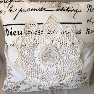 Puede incluir: Cojín decorativo color crema con un gran tapete de ganchillo en forma de estrella. El cojín tiene un estampado floral en la parte inferior y texto escrito en la parte superior. Una rosa de tela está en el centro del tapete.