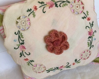 以下が含まれることがあります： フリルのある縁取りと刺繍された花の模様が施された白い枕。枕の中央には、かぎ針編みの花があり、ピンクと白の花柄の生地で囲まれています。