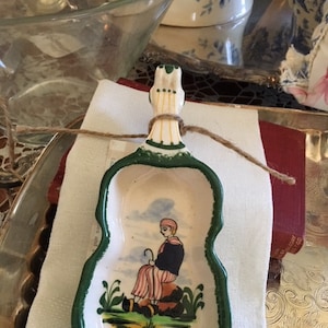 May include: A small, green and white ceramic dish shaped like a violin. The dish features a painted scene of a person in traditional clothing sitting on a rock with a staff.