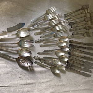 May include: A collection of vintage silverware, including forks, spoons, and a butter knife. The silverware is arranged in a fan shape on a white fabric background.