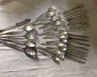 May include: A collection of vintage silverware, including forks, spoons, and a butter knife. The silverware is arranged in a fan shape on a white fabric background.