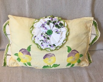 Puede incluir: Una almohada amarilla con un borde de ganchillo y una flor blanca de ganchillo con un botón verde en el centro. La almohada tiene tres pensamientos pintados en morado y amarillo.