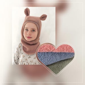 Op de afbeelding: Een kind draagt een bruine fleece muts met aangehechte oren en een bijpassende sjaal. De muts heeft gestreepte details op de oren. Een hartvormige staal toont roze, blauwe en groene fleece stofstalen.