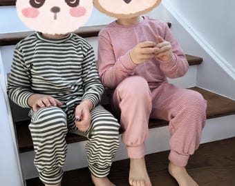 Könnte beinhalten: Zwei Kinder-Pyjama-Sets. Ein Set ist dunkelgrün und weiß gestreift, das andere rosa und weiß gestreift. Beide Sets umfassen langärmlige Oberteile und passende Hosen. Die Kinder sitzen auf Holztreppen.