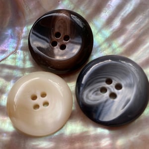 Könnte beinhalten: Drei runde Knöpfe mit jeweils vier Löchern. Die Knöpfe sind aus Perlmutt gefertigt und haben ein Wirbelmuster. Die Knöpfe sind in den Farben Braun, Weiß und Schwarz.