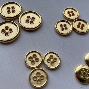 Könnte beinhalten: Zehn goldfarbene Metallknöpfe mit jeweils fünf Löchern. Die Knöpfe sind in einem zufälligen Muster auf einer weißen Oberfläche angeordnet.