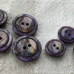 Könnte beinhalten: Eine Sammlung von neun purpurfarbenen und goldenen irisierenden Knöpfen mit jeweils zwei Löchern. Die Knöpfe haben eine glänzende Oberfläche und ein wirbelndes Muster.