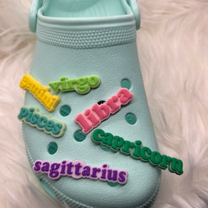 Könnte beinhalten: Ein hellblaues Paar Schuhe mit bunten Charms mit Sternzeichen. Die Charms buchstabieren "Zwillinge", "Jungfrau", "Fische", "Waage", "Steinbock" und "Schütze".