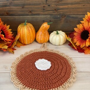 May include: A crocheted pumpkin pie pot holder with a brown crust and a white center. The pot holder is surrounded by fall decor, including pumpkins and sunflowers.