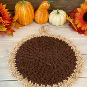 May include: A handmade, round, brown and tan crochet pot holder with a scalloped edge. The pot holder is surrounded by decorative pumpkins and fall foliage, creating a seasonal theme.