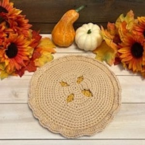 May include: A beige crocheted trivet with a scalloped edge and decorative cutouts, resembling a pie. The trivet is surrounded by fall-themed decorations, including sunflowers, maple leaves, and gourds.